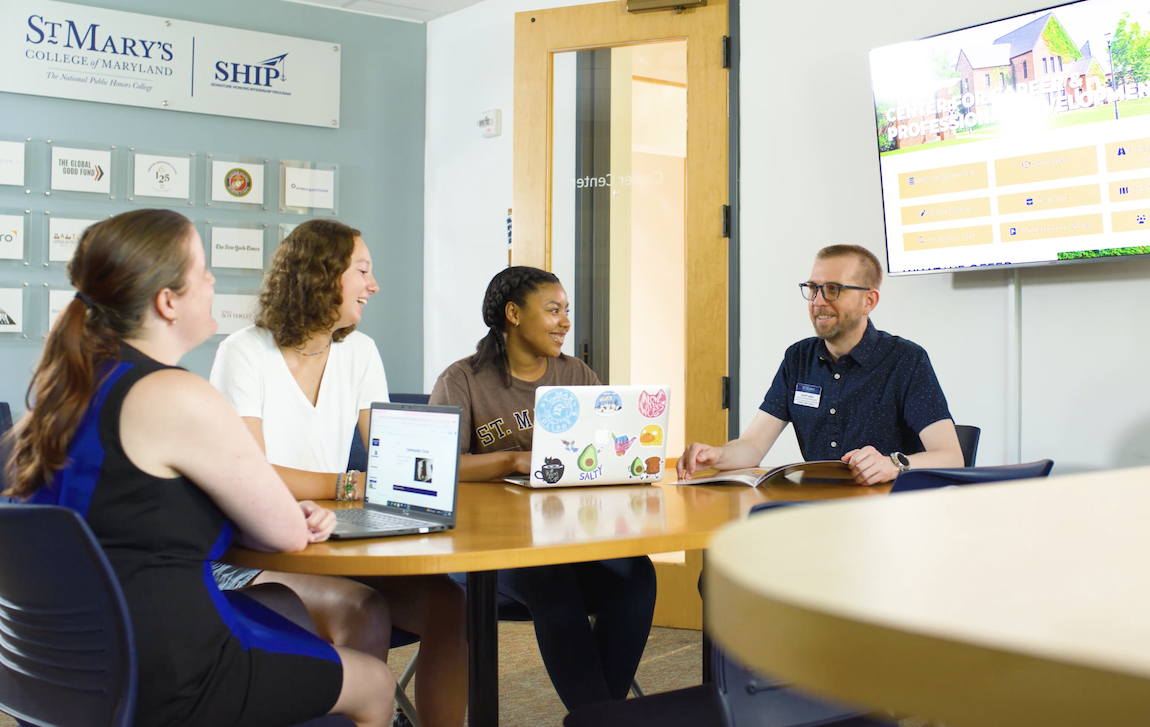Four people sit around a table with laptops, engaged in a discussion. A sign on the wall reads "St. Mary's College of Maryland." A screen displays a colorful website. They appear to be in a meeting room.